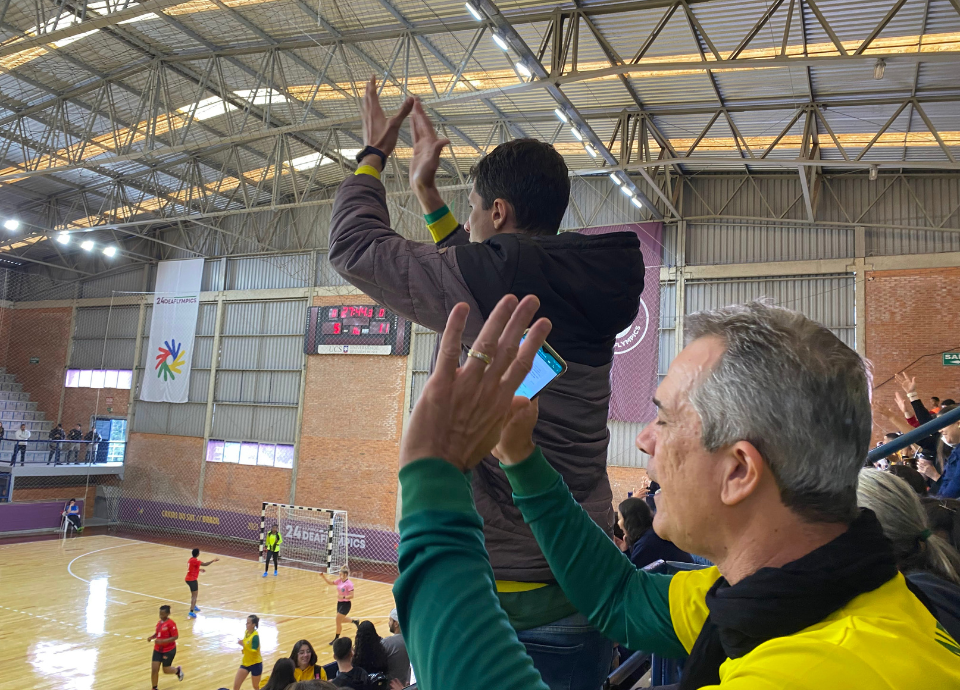 Descrição da imagem: foto do ex-atleta Silvio Caporal e de seu filho Felipe na arquibancada do ginásio de esportes durante a final do Handebol feminino. Eles estão torcendo por Fernanda. Fim da descrição.