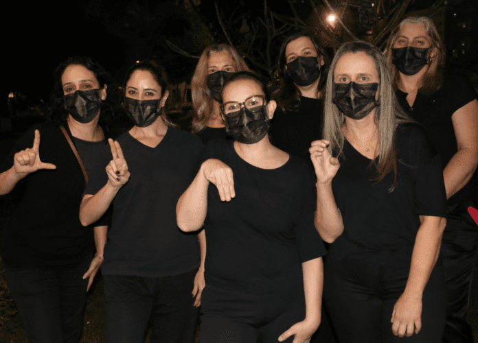 Descrição da imagem: foto de um grupo de mulheres. Todas elas estão vestindo roupas pretas. Elas estão lado a lado e escrevem a palavra LUME com as mãos, sinalizando em Libras. Fim da descrição.