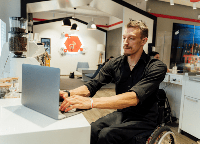 Descrição da imagem: foto de um homem com deficiência física. Ele está em um ambiente corporativo, trabalhando. O homem é cadeirante e está digitando em um notebook. Fim da descrição.