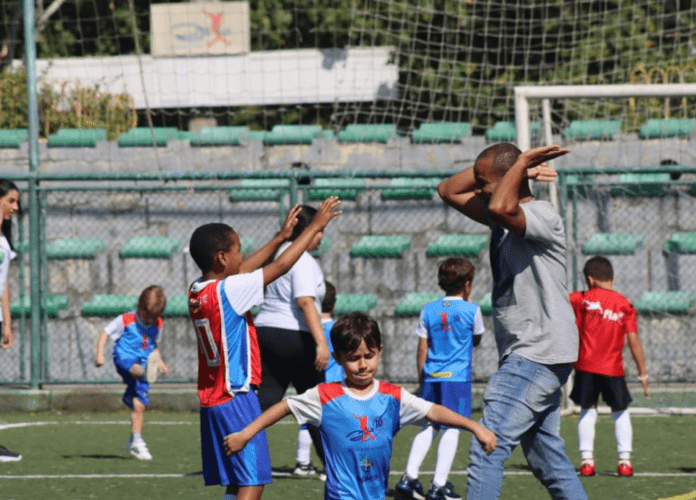 Descrição da imagem: foto da partida de futebol realizada na escolinha Zico 10. Várias crianças estão no campo de futebol. Fim da descrição.