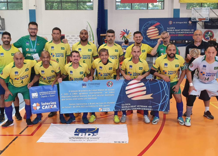 Descrição da imagem: foto dos surdoatletas da Seleção Brasileira Master de Futsal de Surdos. Eles estão perfilados e seguram as bandeiras do Brasil, da DIFA e da CBDS. Fim da descrição.