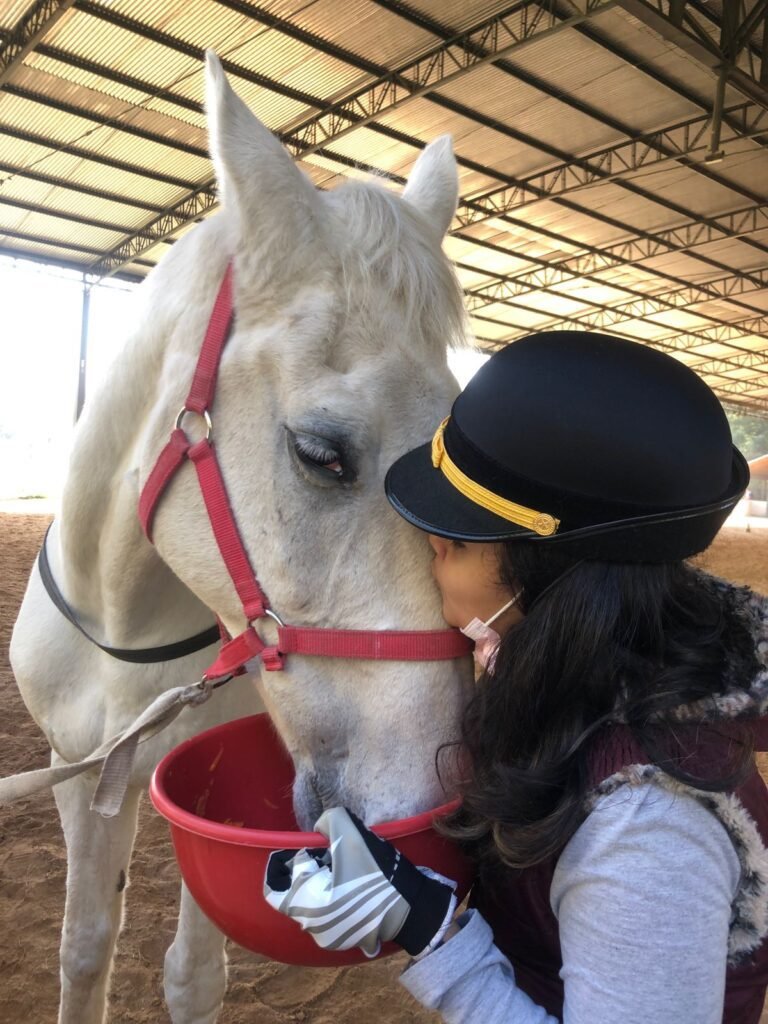 Descrição da imagem: foto de uma profissional de equoterapia beijando um cavalo branco. Fim da descrição.