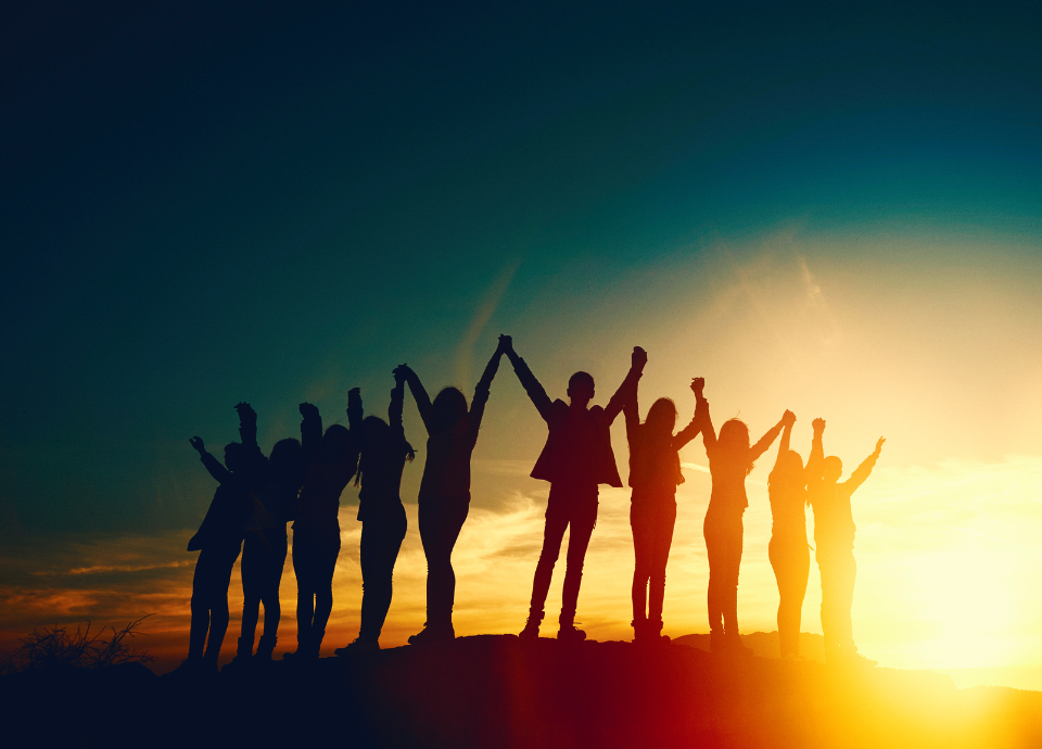 Descrição da imagem: foto de um grupo de jovens lado a lado. Eles estão de mãos dadas e com os braços levantados. Vemos o contorno de seus corpos que estão em contraste ao pôr do sol. Fim da descrição.