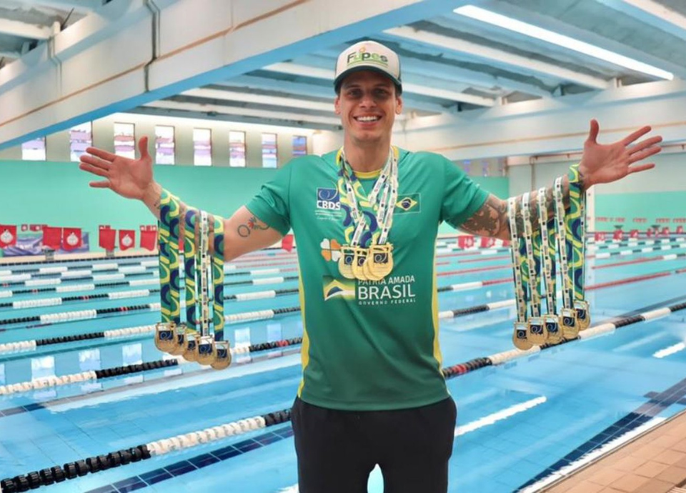 Descrição da imagem: foto do atleta Guilerme Maia. Ele está com os dois braços abertos e exibe neles suas 14 medalhas de ouro. Fim da descrição.