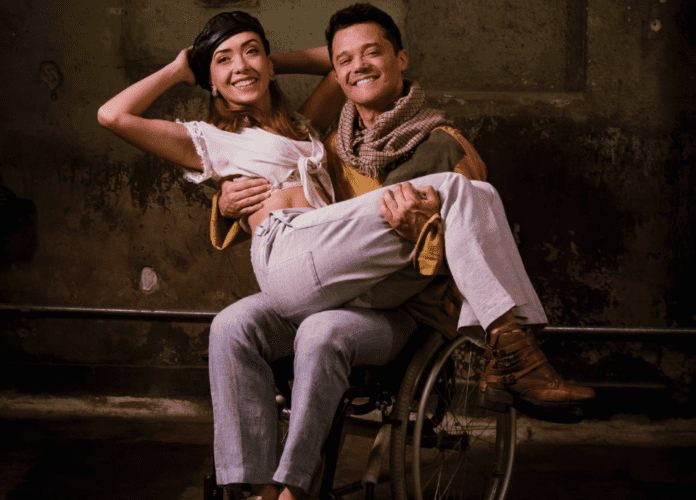 Descrição da imagem: foto de divulgação da peça. Nela estão os atores Ligia e Ubiracy. Ele está sentado em uma cadeira de rodas e segura a atriz em seu colo. Eles estão sorrindo. Fim da descrição.