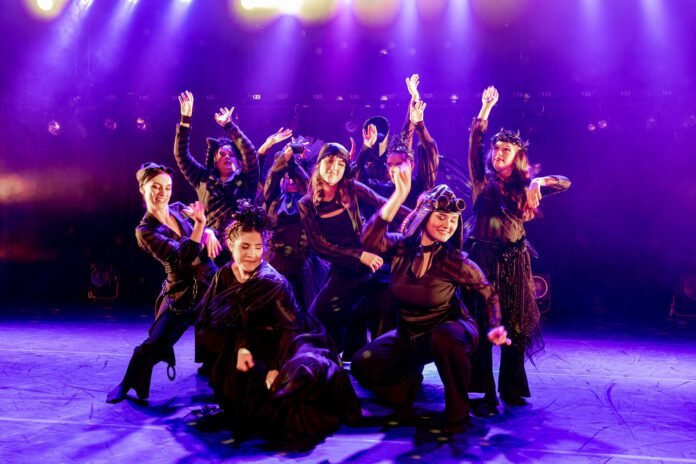 Descrição da imagem: foto de uma cena da peça. Nela estão os atores / bailarinos. O cenário tem fundo preto e o palco está iluminado com uma cor lilás. Fim da descrição.