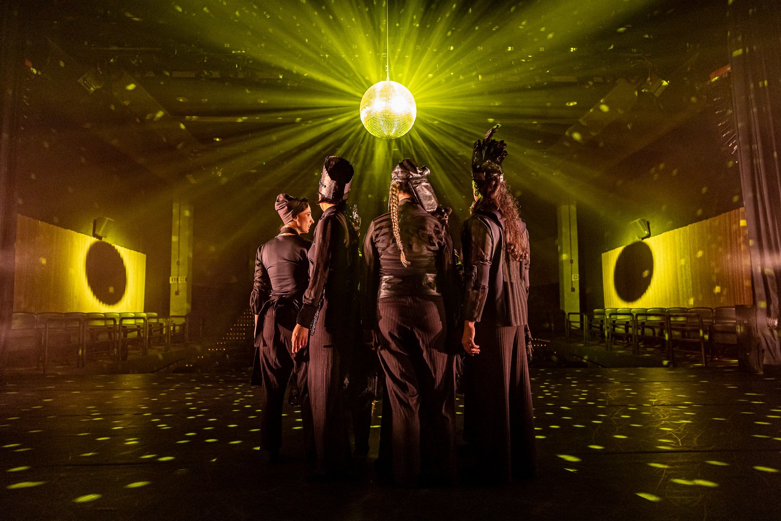 Descrição da imagem: foto de uma cena da peça. Nela estão os atores / bailarinos. Eles fazem a coreografia no centro do palco. Um globo iluminado irradia uma luz amarela em um ambiente escuro. Fim da descrição.