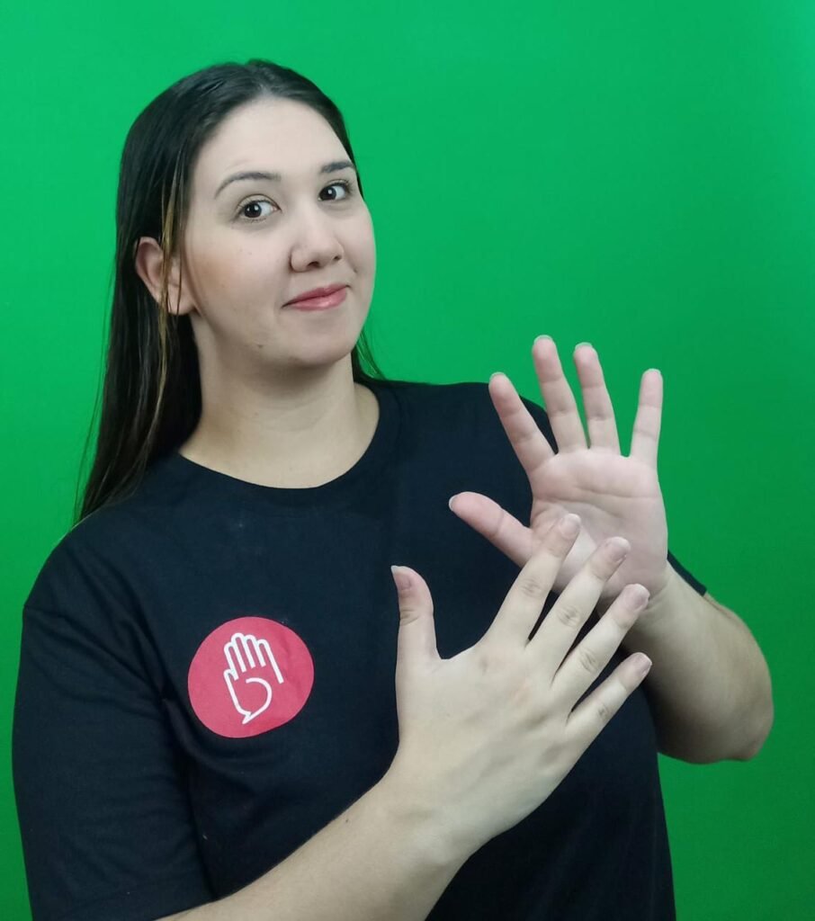 Descrição da imagem: foto da insttutora e intérprete de Libras Cassia. Ela é uma mulher branca, de cabelos castanhos, lisos. Cassia está fazendo uma sinalização em Libras. Fim da descrição.