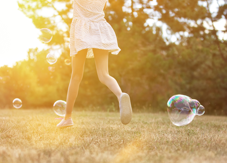 Descrição da imagem: foto de uma menina correndo em um parque. A imagem tem um filtro amarelado e a menina usa um vestido branco e corre entre bolhas de sabão. Fim da descrição.
