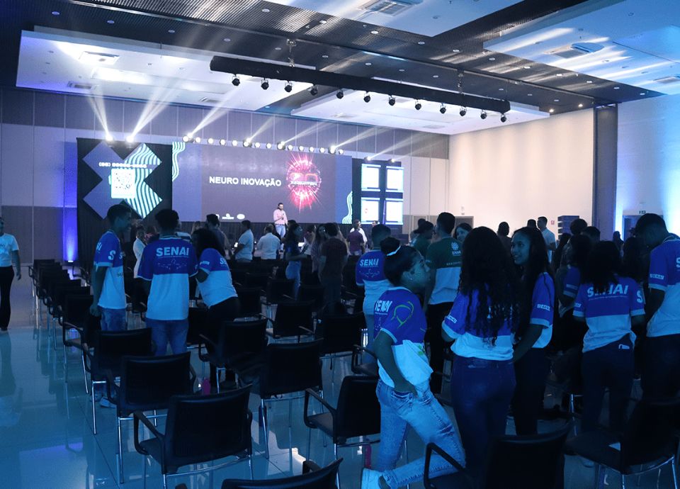 Descrição da imagem: foto do auditório do evento. Várias pessoas estão acompanhando uma apresentação. Fim da descrição.