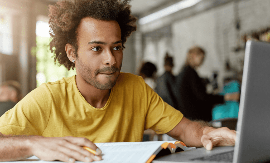 Descrição da imagem: foto de um homem usando um notebook. Ele é negro e está com um livro aberto sobre a mesa. Fim da descrição.