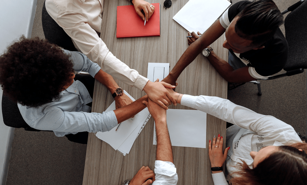 Descrição da imagem: foto de um grupo de pessoas em um ambiente corporativo. Todos estão com as mãos unidas no centro de uma mesa. Fim da descrição.
