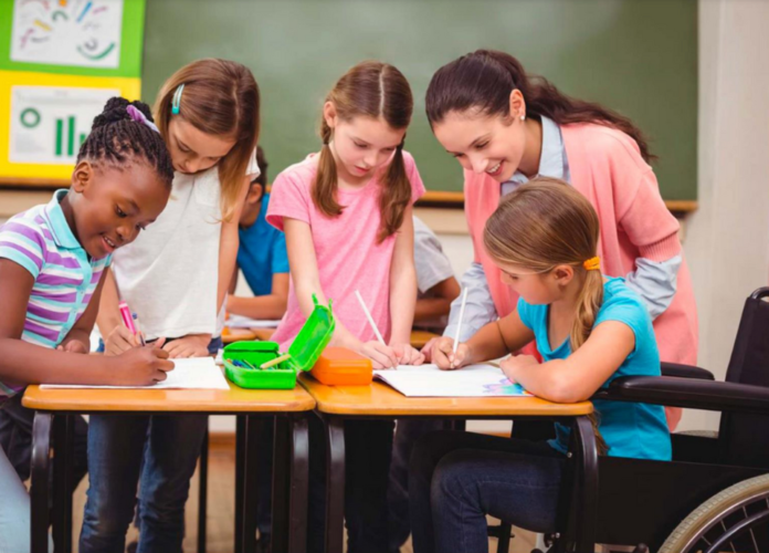 Descrição da imagem: foto de uma sala de aula. Uma educadora e quatro estudantes realizam uma atividade. Eles estão em volta de uma mesa e uma das alunas é cadeirante. Fim da descrição.