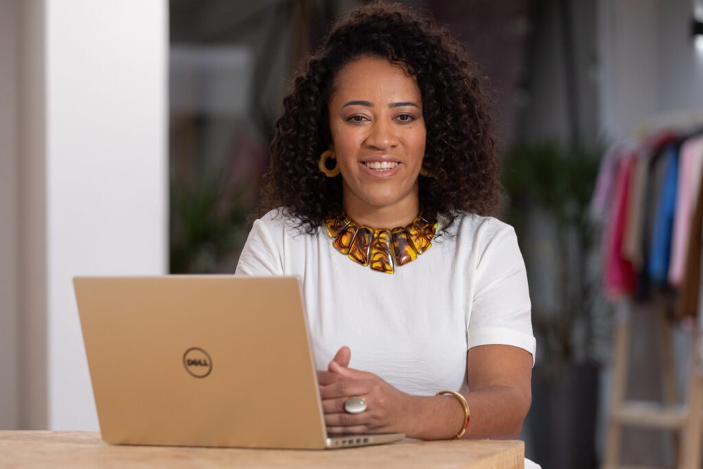 Descrição da imagem: foto de Liliana Rocha. Ela é uma mulher negra, com cabelos castanhos. Liliana está sentada e, à sua frente, tem um notebook aberto. Ela está vestindo uma blusa branca e um colar colorido. Fim da descrição.