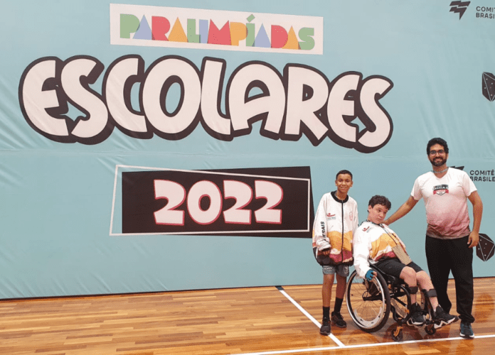 Descrição da imagem: foto do técnico surdo Gabriel com os dois paratletas. Eles estão na frente do banner das Paralimpíadas Escolares 2022. Fim da descrição.