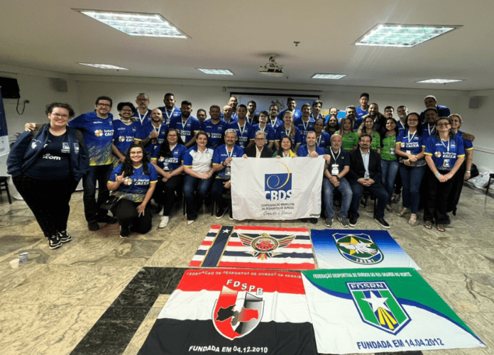 Descrição da imagem: foto de todos os participantes do semiário. No chão, à frente deles, estão bandeiras das associações participantes. Fim da descrição)