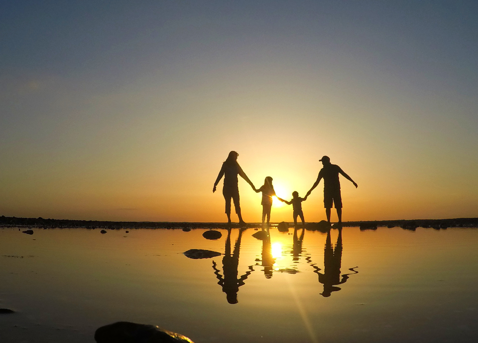 Descrição da imagem: foto de uma família. Pai, mãe e duas crianças, caminhando na beira do mar. Ao fundo vemos o nascer do sol. Fim da descrição