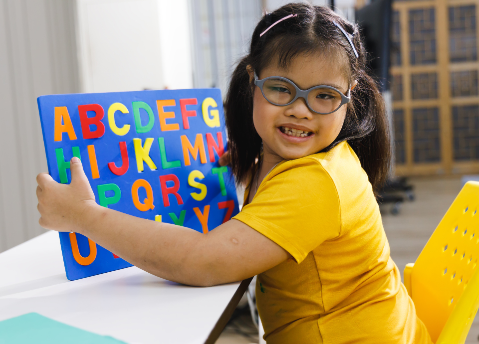 Descrição da imagem: foto de uma menina com síndrome de Down. Ela está segurando uma placa com letras do alfabeto e sorri. Fim da descrição.