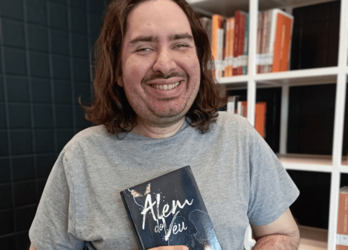 Descrição da imagem: foto do escritor Igor. Ele é um homem branco de cabelos castanhos, lisos, na altura dos ombros. Igor está sorrindo e segura o livro 'Álém do véu'. Fim da descrição.