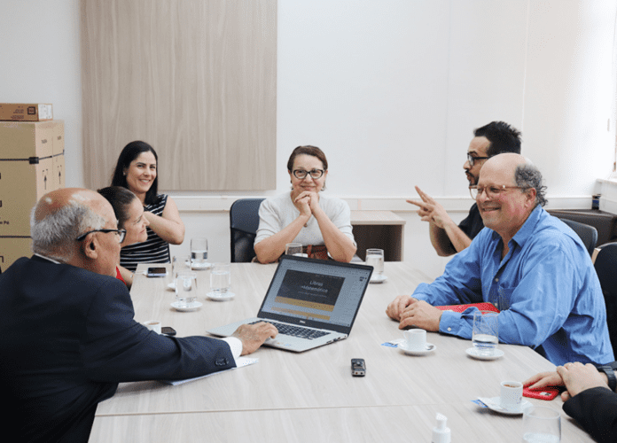 Descrição da imagem: foto de um grupo de pessoas em uma reunião de trabalho. São seis pessoas, sentadas em volta de uma mesa de reuniões. Sobre a mesa estão notebooks e xícaras de café. As pessoas estão conversando. Fim da descrição.
