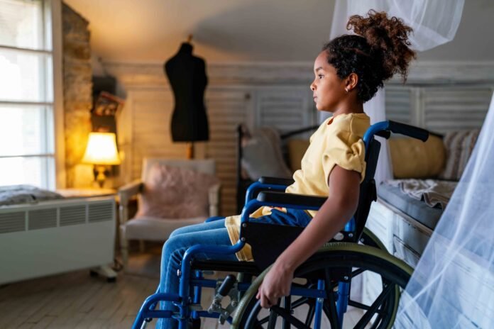 Descrição da imagem: foto de uma menina cadeirante. A menina está em um quarto. Fim da descrição.