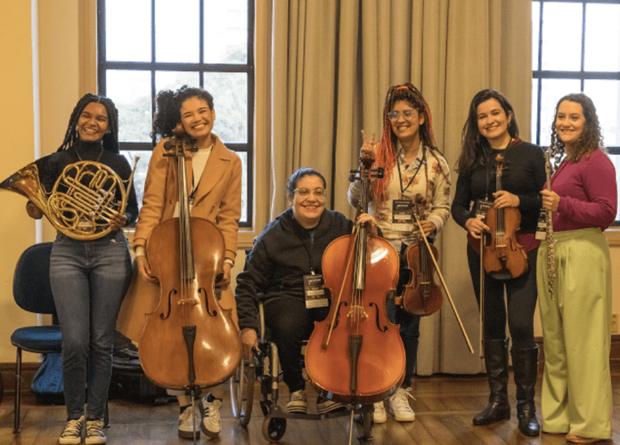 Descrição da imagem: foto das integrantes da 1ª orquestra parassinfônica do Brasil. São seis mulheres que estão lado a lado, acompanhadas de seus instrumentos musicais. Fim da descrição.