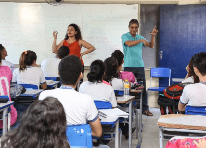 Descrição da imagem: foto de uma sala de aula. Alunos estão sentados em suas carteiras. Na frente da sala estão a professora e o intérprete de Libras. Fim da descrição.