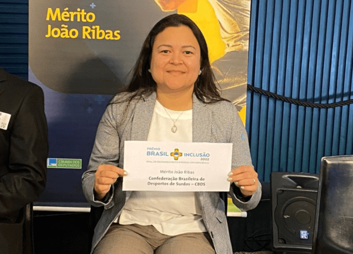 Descrição da imagem: foto de Diana Kyosen com o certificado do prêmio. Diana é uma mulher com traços orientais e tem cabelos castanhos, lisos e compridos. Ela está vestindo uma calça cáqui, uma blusa branca e um terninho cinza. Fim da descrição.
