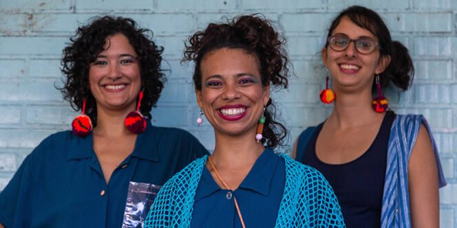Descrição da imagem: foto de três mulheres. Uma delas está na frente e as outras duas estão atrás dela, nas laterais. Elas estão sorrindo. Fim da descrição.