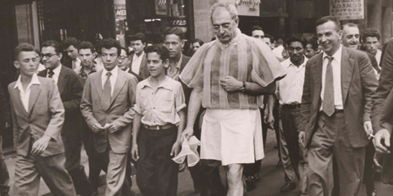 Descrição da imagem: foto em preto e branco de várias pessoas caminhando em uma rua. Fim da descrição.