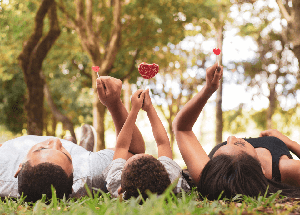 Descrição da imagem: foto de uma família, pai, mãe e filha. Eles estão deitados na grama, em um parque, e seguram pirulitos em forma de coração. Fim da descrição.