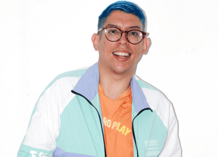 Descrição da imagem: foto do influenciador Ivan Baron. Ele está vestindo uma camiseta na cor laranja em blusão colorido nas cores verde, azul e branco. Ivan tem cabelos curtos e lisos, que estão pintados de azul. Ele usa óculos de grau. Fim da descrição.