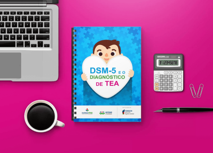 Descrição da imagem: Foto de uma mesa vista de cima. A mesa é pink e sobre ela estão um notebook, uma xícara de café, uma calculadora e uma caneta, além da cartilha 'DSM-5 e o diagnóstico de TEA', que está o centro. Fim da descrição.