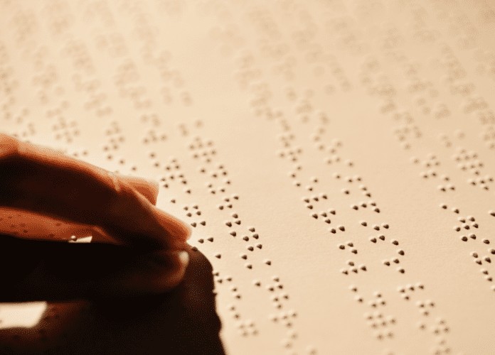 Descrição da imagem: foto de dedos fazendo a leitura em braille em um documento impresso. Fim da descrição.