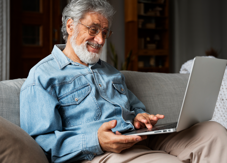 Descrição da imagem: foto de um homem sentado no sofá, usando um notebook. Ele tem cabelos e barbas grisalhos, e está vestindo uma camisa jeans e uma calça bege. Fim da descrição.