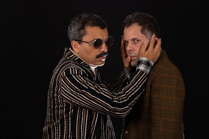 Descrição da imagem: foto de uma cena da peça. Nela estão os dois atores que encenam a peça. O ator que interpreta um cego está tocando o rosto do outro ator. Fim da descrição.