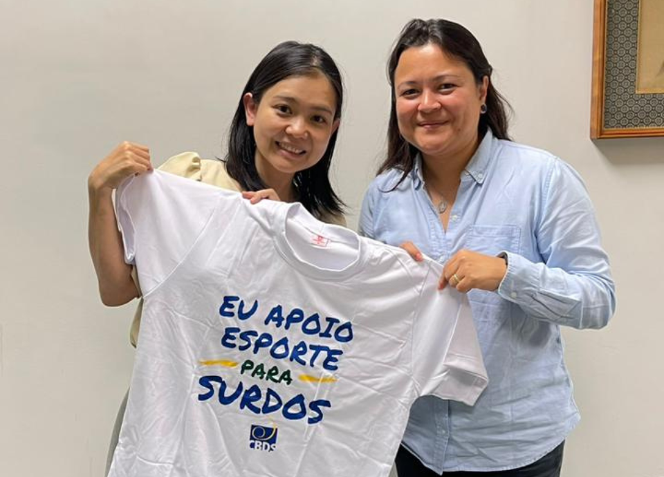 Descrição da imagem: foto de Diana com a vice-cônsul do Japão, Akiko Kikuchi. Elas estão segurando uma camiseta branca com a frase: eu apoio o esporte para surdos. Fim da descrição.