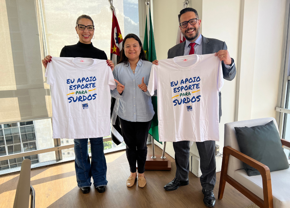 Descrição da imagem: Diana com a secretária-adjunta da SMPED, Dika Vidal e o chefe de gabinete da SEME, Ricardo Calciolari. Eles estão segurando uma camiseta branca com a frase: eu apoio o esporte para surdos. Fim da descrição.