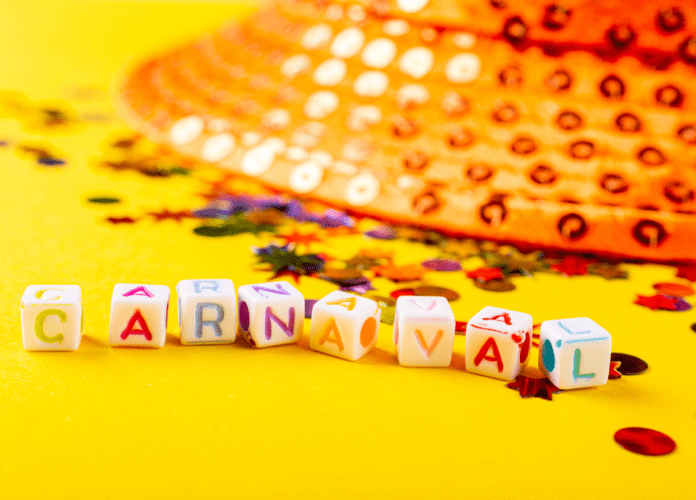 Descrição da imagem: foto com objetos que remetem ao Carnaval. Em primeiro plano está uma sequência de pequenos cubos com letras coloridas que formam a palavra CARNAVAL. Em segundo plano está um chapeu com lantejoulas, que também estão espalhadas à sua volta. Os objetos estão sobre uma base amarela. Fim da descrição.