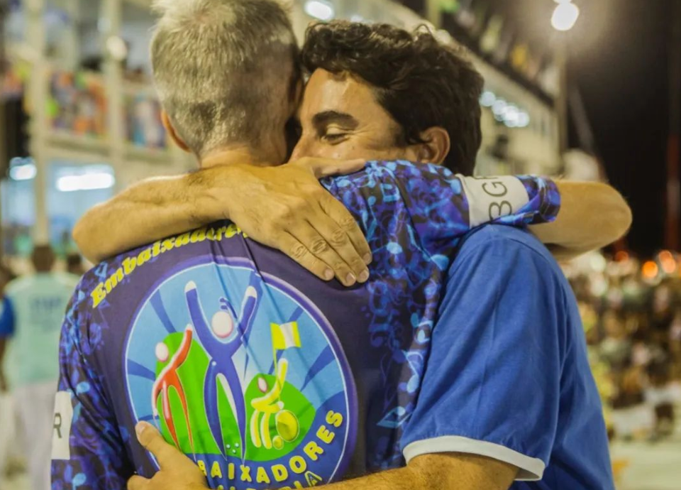 Foto de Paul e Caio se abraçando. Ao fundo está a arquibancada da Marquês de Sapucaí. Fim da descrição.