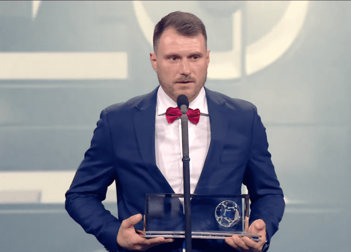 Imagem de Oleksy durante o discurso de agradecimento. Ele está segurando o prêmio. Fim da descrição.