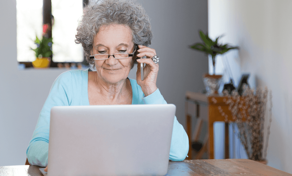 #descricaodaimagem foto de uma mulher idosa usando um computador. Ela está falando no celular. Fim da descrição.