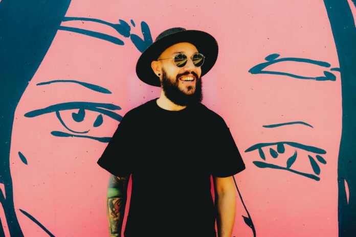 #descricaodaimagem foto do DJ Robbie Wilde. Ele é um homem branco com barba. Robbie usa uma camiseta preta, óculos escuros e chapéu preto. Fim da descrição.