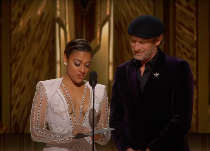 #descricaodaimagem imagem dos atores Ariana DeBose e Troy Kotsur no palco da cerimônia do Oscar, no momento da abertura do envelope com o nome do premiado. Fim da descrição.