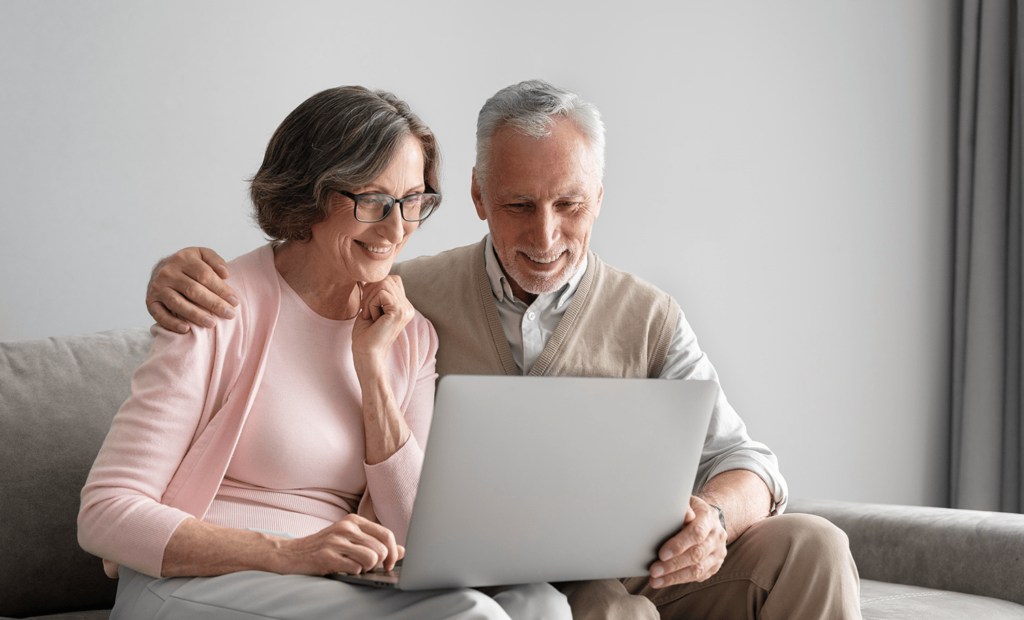 #descricaodaimagem foto de uma mulher e um homem idosos, sentados lado a lado, usando um notebook. Fim da descrição.
