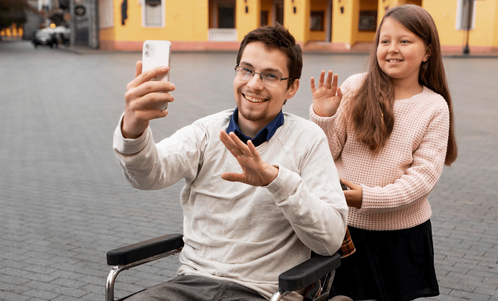 #descricaodaimagem #pratodosverem foto de duas pessoas na rua sorrindo para um foto, uma delas é um homem branco, cadeirante que segura o celular e tira foto e atrás dele temos uma menina branca, com cabelos longos, acenando para foto.