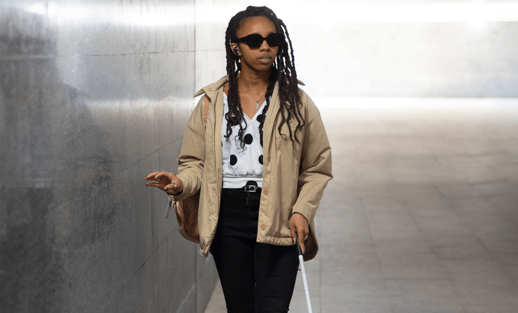 #descricaodaimagem #pratodosverem foto de uma mulher cega. Ela é negra, tem tranças, usa óculos escuros e uma bengala. Ela está andando por um corredor.