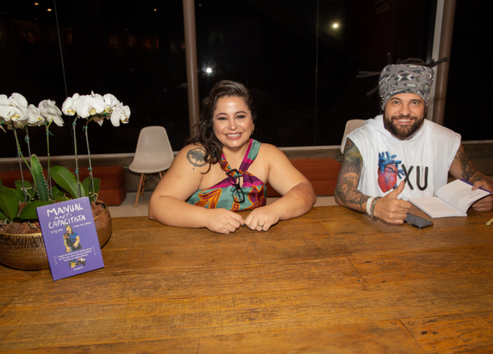 #descricaodaimagem Foto de Carolina e Billy durante o lançamento do livro. Eles estão lado a lado e, ao lado, está o livro, em destaque, apoiado em um vaso de orquídeas. Fim da descrição.