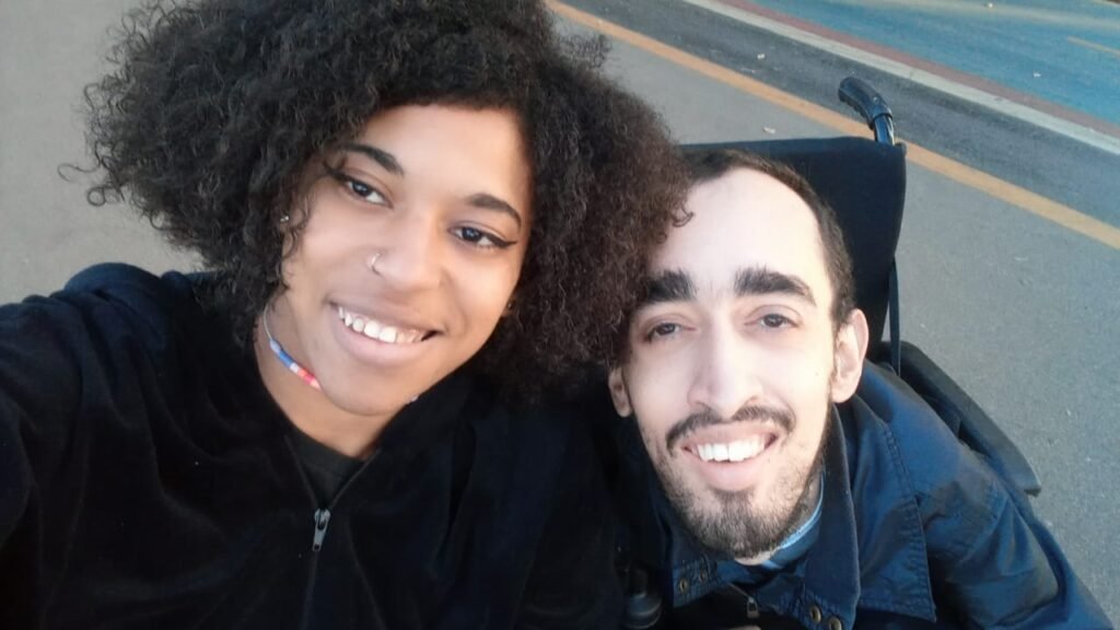 #descricaodaimagem #pratodosverem foto dos namorados Samantha e Felipe. Samantha é uma mulher negra de cabelos cacheados, na altura dos ombros. Felipe é um homem branco, cadeirante, com cabelos e barba castanhos. Eles estão lado a lado e sorriem.