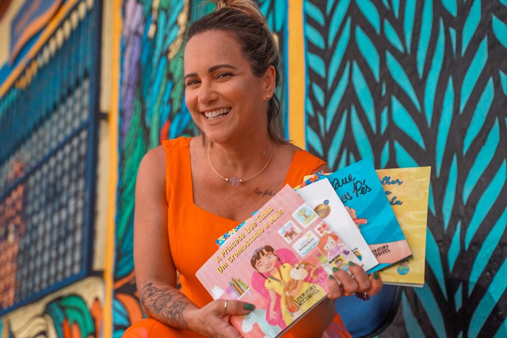 #descricaodaimagem #pratodosverem foto de Carina Alves. Ela é uma mulher branca de cabelos castanhos que estão presos. Carine está vestindo uma roupa laranja e segura os livros da coleção Literatura Acessível. Ela sorri.